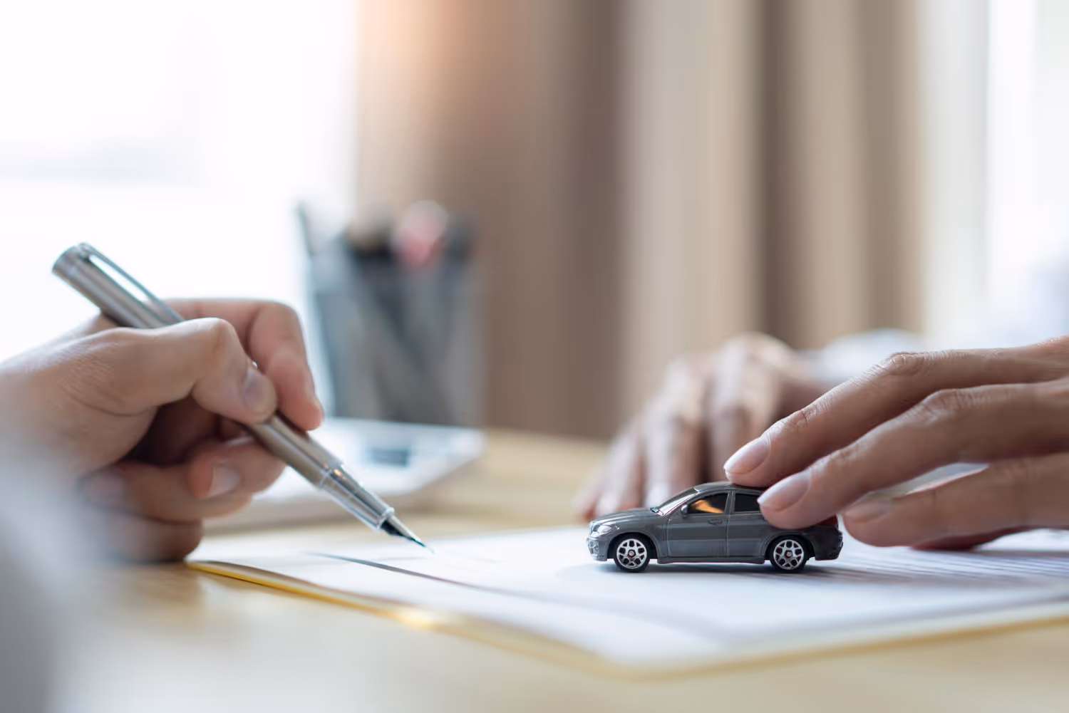Close-up of a hand holding a pen signing a document and another hand touching a small toy car on the paper.