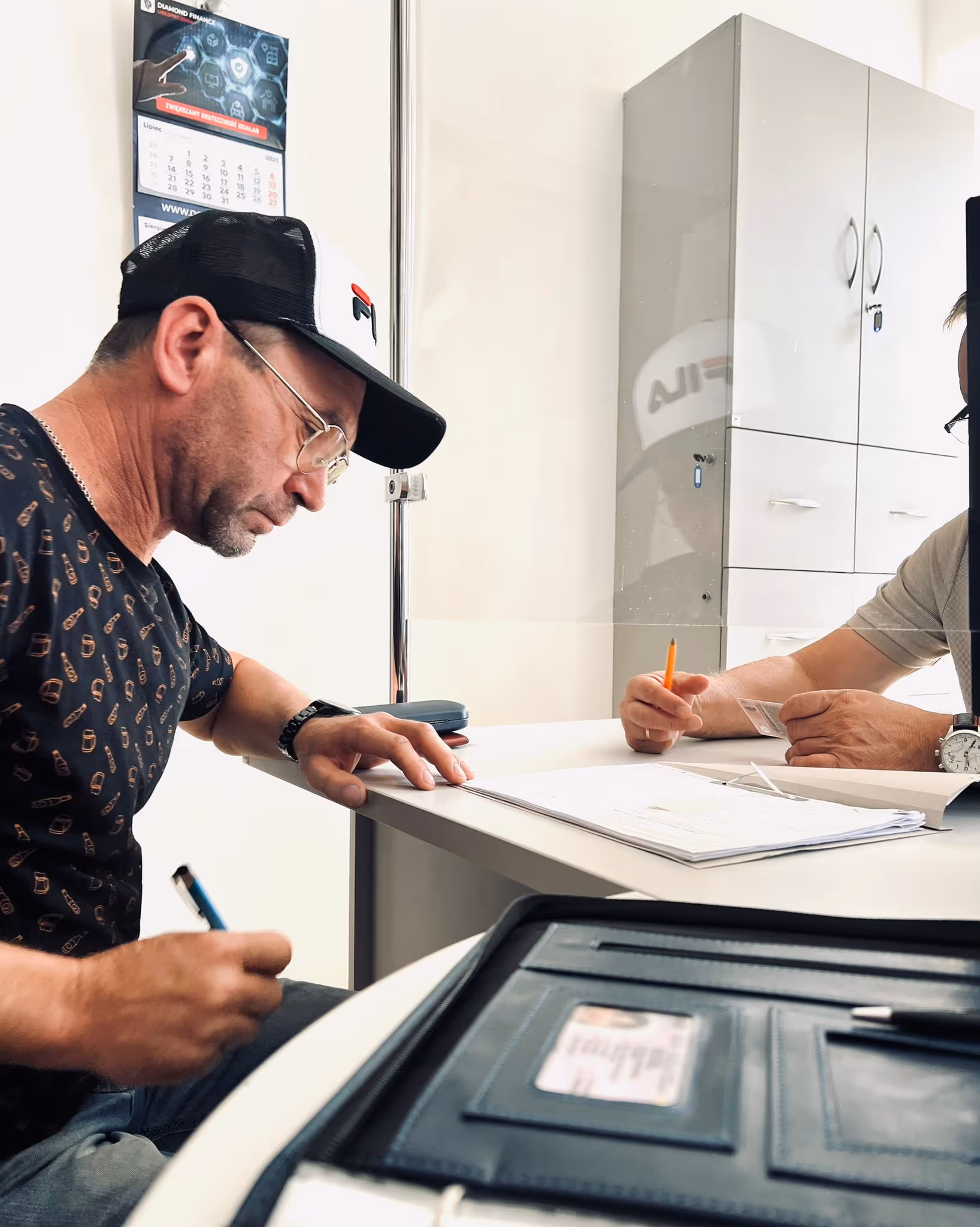 Person in green sweatshirt holding a Polish ID card across a desk with a transparent barrier, another person partially visible on the other side.